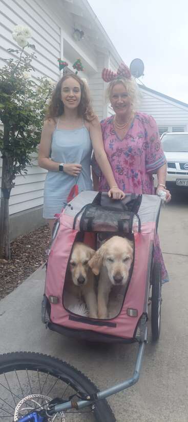 Two women in festive headbands smile outside a house. They stand beside a pink dog stroller holding two golden retrievers. A bicycle is attached in front.