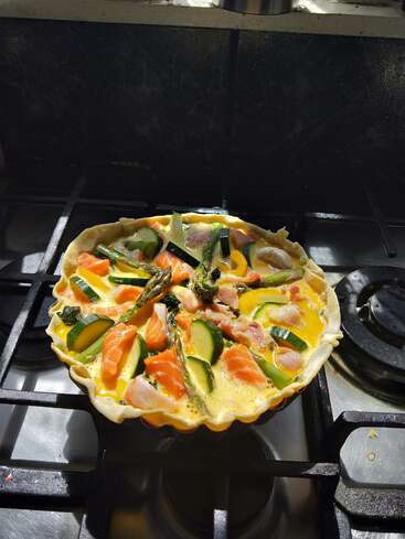 En un molde para tartas hay una colorida quiche cruda rellena de huevos, salmón, calabacín, espárragos y beicon, tomando el sol sobre un hornillo.