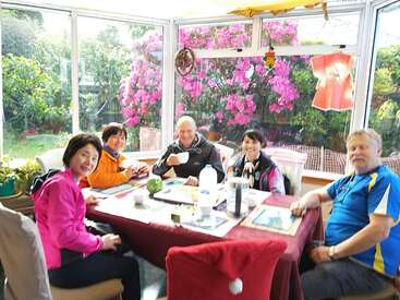 Cinco personas se sientan alrededor de una mesa de comedor en una terraza acristalada, disfrutando de las bebidas y de la compañía de los demás. En el exterior florecen brillantes flores rosas que crean un ambiente alegre.