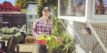 Un hombre sonriente con camisa de cuadros y gorra está de pie en un jardín soleado, sosteniendo una cesta de verduras recién recogidas junto a una casa con un cartel de "Bienvenido".