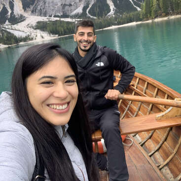 Das Bild zeigt ein Selfie von einer Frau und einem Mann in einem Holzboot auf einem See, mit Bergen und Bäumen im Hintergrund.
