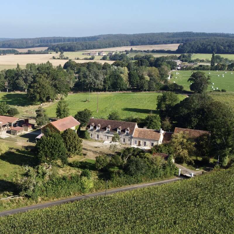 La imagen representa un sereno paisaje rural con una gran casa, graneros y campos, con colinas onduladas y árboles de fondo bajo un cielo azul despejado.