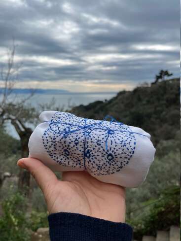 Eine Hand hält ein kleines, gefaltetes weißes Tuch mit verschlungenen blauen Mustern vor einem malerischen Hintergrund aus Hügeln, Bäumen und einem bewölkten Himmel in der Nähe von Wasser.