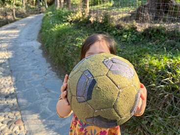 Ein Kind hält einen abgenutzten, gelben Fußball in die Kamera, der sein Gesicht verdeckt. Ein sonnenbeschienener Weg und Grünpflanzen schaffen eine friedliche Umgebung.