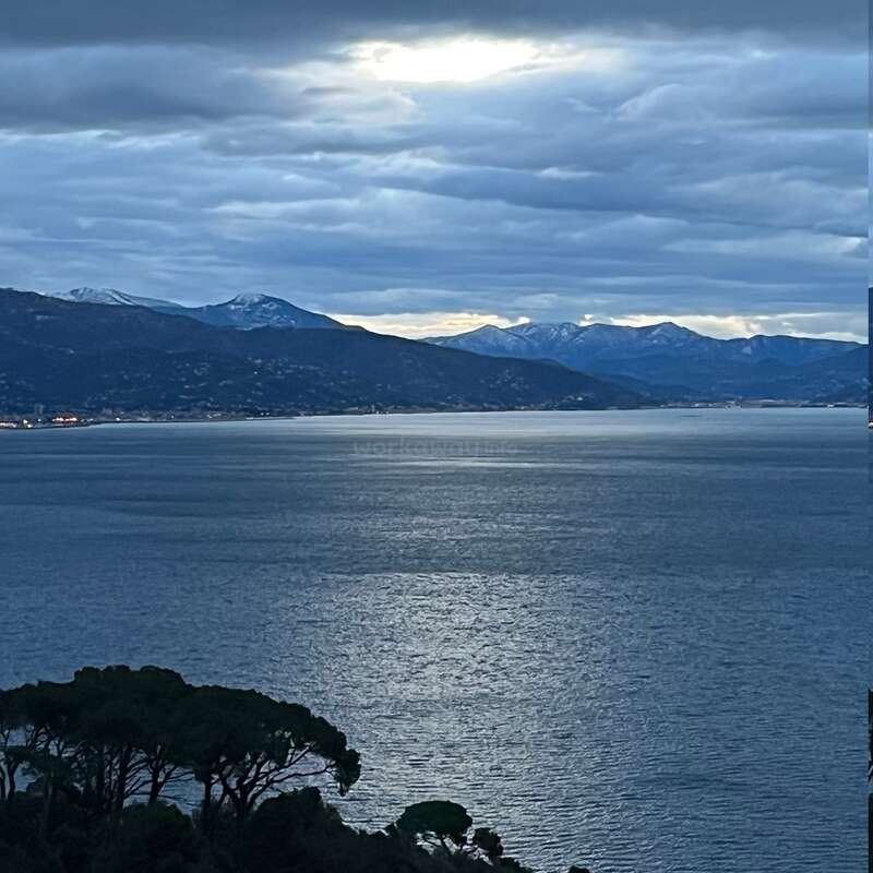 Bewölkter Himmel über einem friedlichen See, durch den das Sonnenlicht bricht. Die Berge in der Ferne sind leicht schneebedeckt. Silhouettierte Bäume umrahmen den Vordergrund und schaffen eine heitere, ruhige Szene.