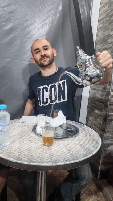 A man at a round table pours tea from a metal teapot into a glass. He wears an "ICON" shirt. Water, sugar, tissue visible.