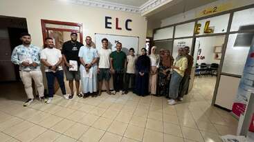 A group of thirteen people stands smiling in a well-lit room with "ELC" on the wall, suggesting a formal gathering or educational event at a learning center.