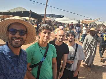 Four friends pose for a selfie at a busy outdoor market. Sun hats, smiles, market stalls, and diverse shoppers create a lively, colorful, and sunny scene.