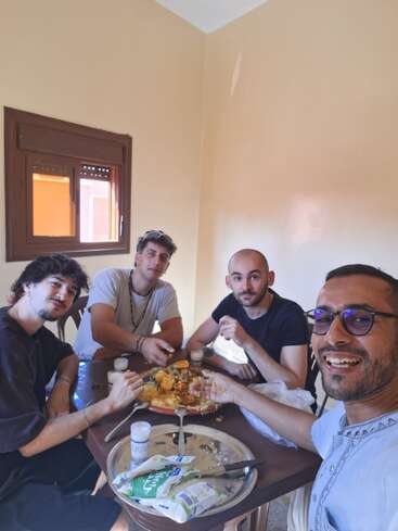 Four friends sit around a table, smiling and enjoying a shared meal. Natural light enters through the window, creating a warm, friendly, and relaxed atmosphere.