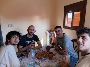Four young men sit around a table, sharing a meal together. They are smiling, with a dish of food, cups, and water on the table.