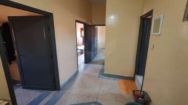 Cette image montre un couloir avec des murs beiges et plusieurs portes sombres ouvertes menant à différentes pièces. Le sol est orné de motifs et d'une corbeille à papier.