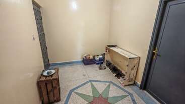 Une pièce simple aux murs crème est dotée d'un sol à motifs. Des chaussures sont rangées dans un coin, des vivres et des caisses à proximité, et un coffre en bois est présent.