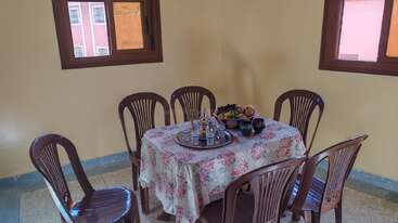 Un petit coin repas avec six chaises en plastique marron entoure une table carrée recouverte d'une nappe fleurie. Des fruits et un service à thé traditionnel sont disposés avec soin.