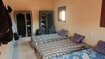 Cette image montre une chambre simple avec trois lits simples, une literie à motifs, deux fenêtres, des armoires en tissu, des crochets muraux et des sacs à dos, créant ainsi une atmosphère de dortoir.