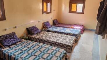 Cette image montre une chambre simple et propre avec quatre lits simples alignés contre le mur. Chaque lit est équipé d'un oreiller et d'une couverture, et il y a deux fenêtres.