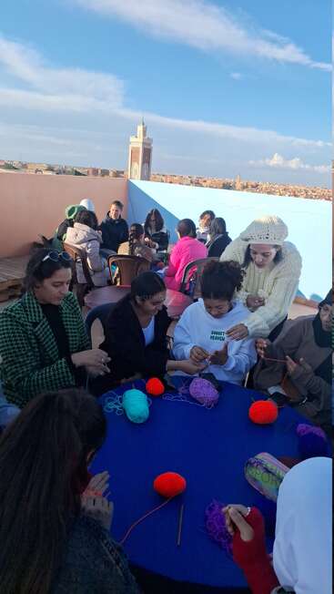 Un groupe de personnes assises autour de tables sur un toit, tricotant avec des fils colorés. L'arrière-plan montre un paysage urbain, un ciel bleu et un minaret.