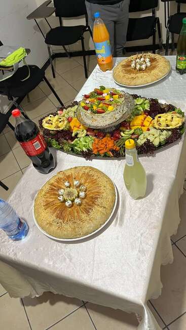 Une table de fête garnie de deux grandes tartes, d'un plateau de salades éclatantes, de boissons non alcoolisées et d'eau, prête pour un repas de fête dans une pièce confortable.