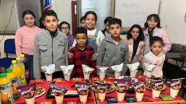 Un groupe d'enfants souriants se tient derrière une table remplie de snacks et de boissons, célébrant un anniversaire. Un tableau blanc à l'arrière-plan indique "Joyeux anniversaire".