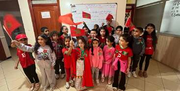 Un groupe d'enfants joyeux se tient ensemble à l'intérieur, tenant et agitant des drapeaux marocains, vêtus de vêtements de fête, célébrant un événement national ou culturel à l'école.