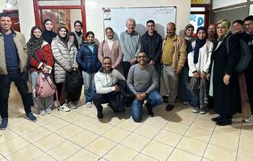 Un groupe de personnes souriantes, hommes et femmes, pose à l'intérieur devant un tableau blanc. Les lettres "ELC" sont affichées sur le mur au-dessus d'eux.
