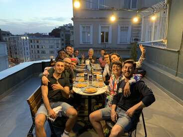 The image depicts a group of people gathered around a table on a rooftop patio, enjoying food and drinks, with a cityscape visible in the background.