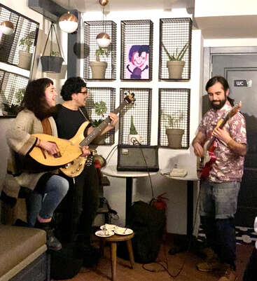 The image depicts three musicians playing guitars in a cozy room with plants and artwork on the walls, surrounded by a warm and intimate atmosphere.