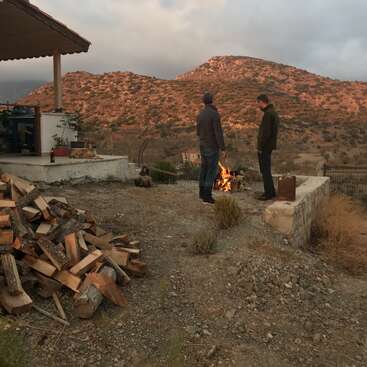 Dois homens estão perto de uma fogueira em um quintal com uma pilha de lenha, cercado por uma casa e uma paisagem montanhosa sob um céu nublado.