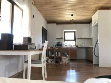 This image shows a cozy kitchen and dining area with wooden floors, white cabinets, a bookshelf, record player, large window, and a wooden ceiling with natural light.
