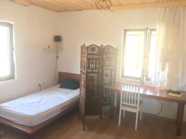 This image shows a simple, sunlit bedroom with a single bed, wooden desk, white chair, ornate room divider, two windows, wooden ceiling, and minimal decor.