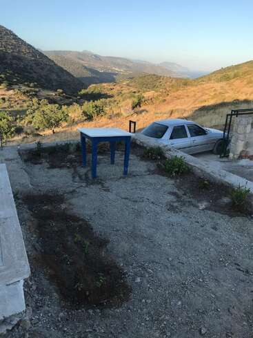 A blue table stands on a patio overlooking dry hills and distant mountains. A white car is parked nearby, with the sea visible far away.