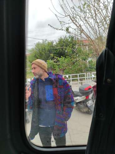 A man wearing a colorful patterned jacket and a beanie stands outside, viewed through a window. He is near a white fence and a parked scooter.