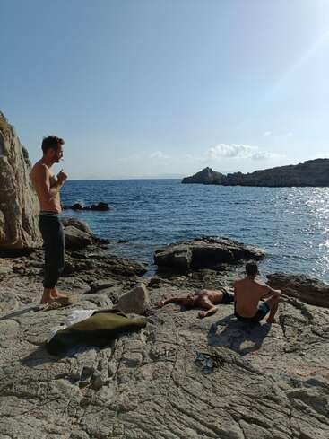 Three men relax shirtless on rocky seaside cliffs under a clear blue sky, surrounded by sparkling ocean water and rugged coastline, enjoying a peaceful, sunny day.