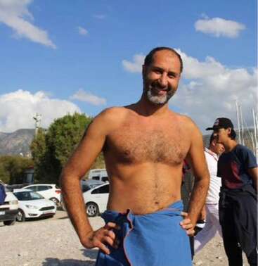 A smiling, shirtless man stands outdoors with his hands on his hips, wrapped in a blue towel. Cars, people, trees, mountains, and a blue sky in background.