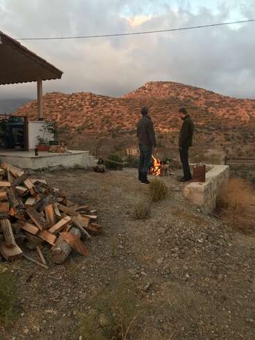Deux hommes se tiennent près d'un foyer dans une cour avec un tas de bois, entourée d'une maison et d'un paysage montagneux sous un ciel nuageux.