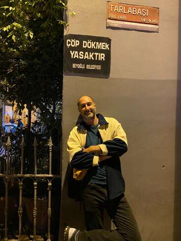 A man leans casually against a corner wall under two signs at night. One sign warns against littering; the other marks "Tarlabaşı Boulevard" in Turkish.