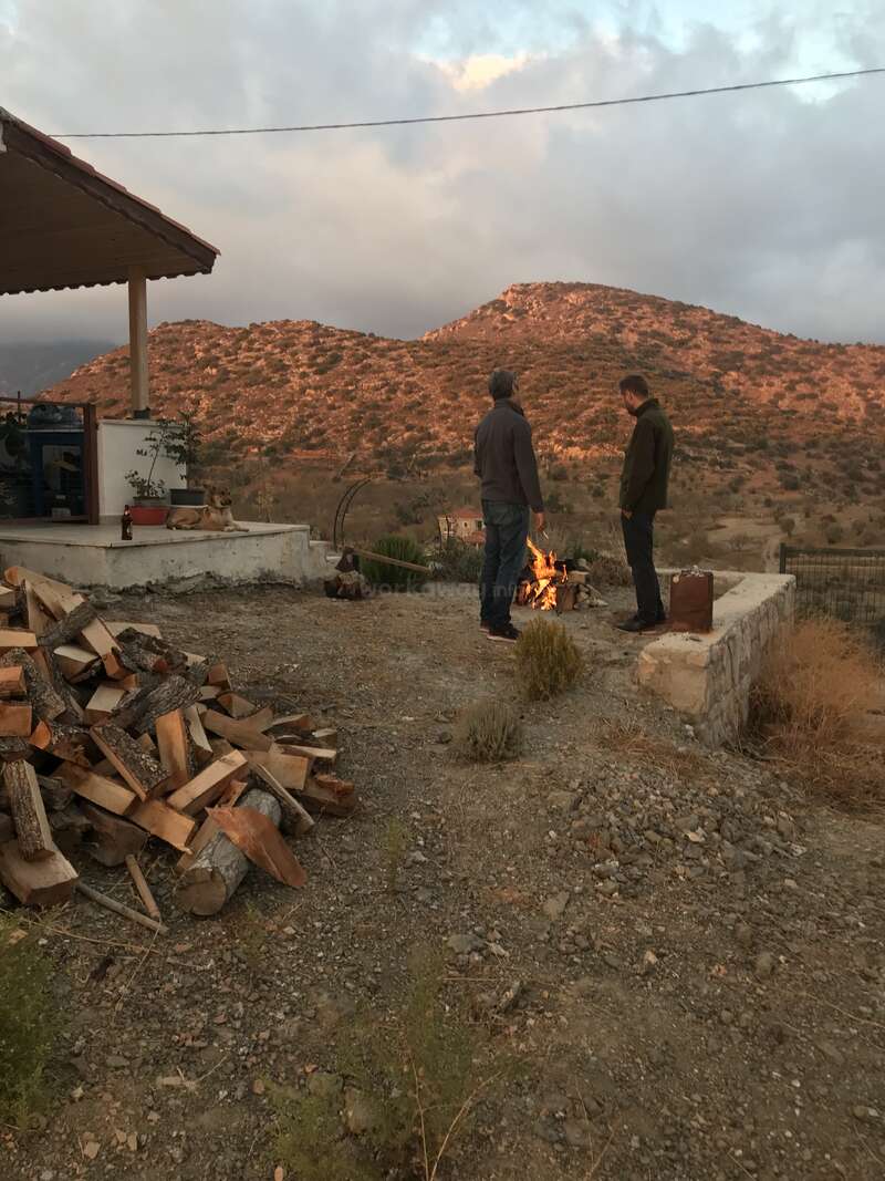 Two men stand near a fire pit in a yard with a woodpile, surrounded by a house and a mountainous landscape under a cloudy sky.