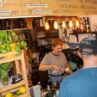 Un barman souriant sert des boissons derrière un comptoir confortable et bien éclairé. Des herbes fraîches et des citrons décorent le comptoir. Les clients discutent, créant une atmosphère animée et accueillante.
