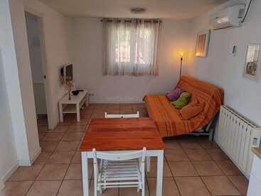 A cozy, minimalist living room with a small wooden dining table, white chairs, an orange sofa bed, wall art, TV, tiled floor, and soft natural light.