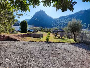 This image shows a peaceful countryside scene with a gravel yard, garden, trees, rustic fences, distant white house, mountain backdrop, and a sunny blue sky.