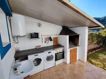 This image shows an outdoor kitchen with a washing machine, dryer, oven, sink, microwave, water heater, countertop, utensils, and a barbecue grill under a slanted roof.