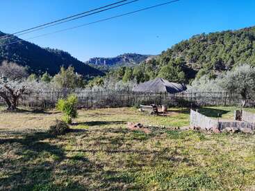 A serene rural landscape with lush green hills, a bamboo fence, small thatched hut, scattered trees, a bench, clear blue sky, and power lines overhead.