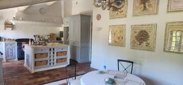 La imagen muestra una cocina con mesa y sillas, dotada de una gran isla con fregadero y fogones, acompañada de una zona de comedor con mesa y sillas. La estancia está bien iluminada y tiene una estética moderna.