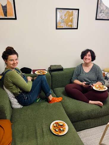 Deux femmes sont assises sur un canapé vert, chacune tenant une assiette de nourriture, et sourient. Des œuvres d'art sont accrochées au mur au-dessus d'elles. L'atmosphère est chaleureuse et conviviale.