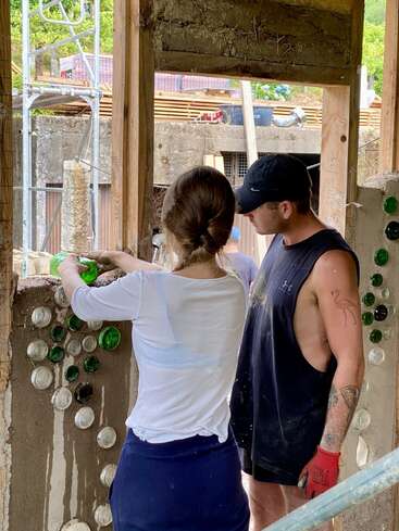Dos personas construyen un muro con botellas de vidrio recicladas. Trabajan juntos, colocando las botellas en cemento húmedo, creando un diseño de construcción único y ecológico. Escenario al aire libre.