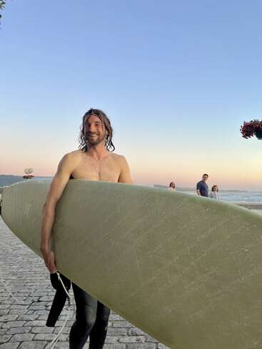 Un hombre sonriente con una gran tabla de surf de pie en un camino empedrado junto a la playa al atardecer, con otros y el océano visible en el fondo.