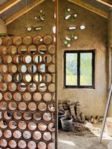 Esta imagen muestra una casa respetuosa con la tierra, con tabiques circulares de tubos de arcilla, paredes enlucidas de barro, ventanas de botellas de vidrio, luz solar, vigas de madera y materiales de construcción apilados en el interior.