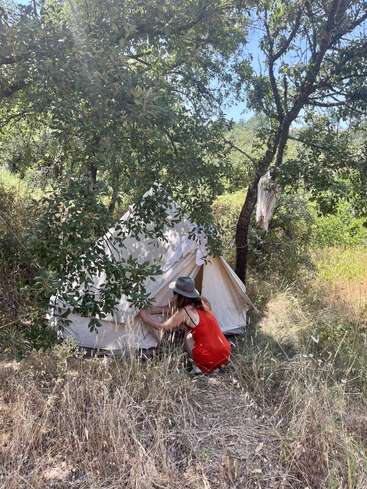 Una mujer con vestido y sombrero rojos se agazapa fuera de una tienda de campaña en una zona soleada y arbolada, rodeada de árboles, hierba y luz natural difusa.