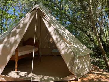 Una tienda de lona se alza sobre una plataforma de madera en medio de los árboles. La luz del sol se cuela entre las hojas, proyectando sombras. En el interior hay una cama de madera y algunos muebles básicos.