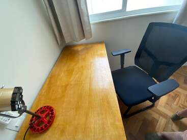 A imagem mostra uma mesa de madeira ao lado de uma janela com as cortinas fechadas, uma cadeira de escritório de malha preta, um abajur vermelho bem diferente e parte de um piso de madeira.