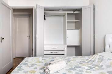 Um quarto limpo e minimalista tem um armário aberto com prateleiras e gavetas, portas cinza-claro, roupa de cama suave com um padrão de folhas e toalhas bem dobradas sobre a cama.